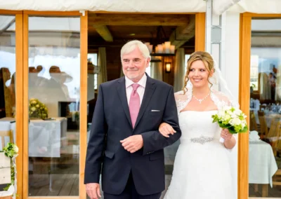 Ein älterer Mann im Anzug führt eine lächelnde Braut in einem weißen Kleid mit Brautstrauß an seinem Arm.