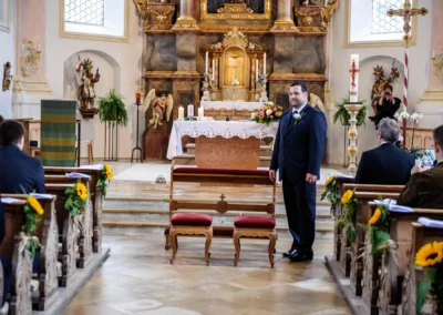 Ein Bräutigam steht vorne in einer geschmückten Kirche, eingefasst von holzgeschnitzten Bänken mit Sonnenblumen.