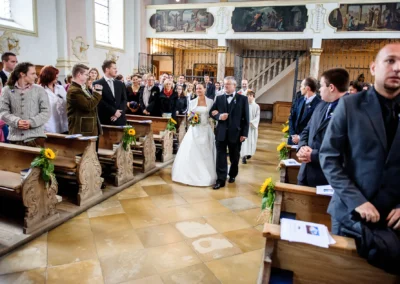 Eine Braut in weißem Kleid schreitet mit einem älteren Herrn den Kirchgang entlang, umgeben von stehenden Gästen.