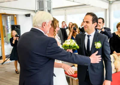 Ein Mann übergibt die Braut mit Blumenstrauß an den Bräutigam. Gäste mit Kameras im Hintergrund. Feierliche Stimmung.