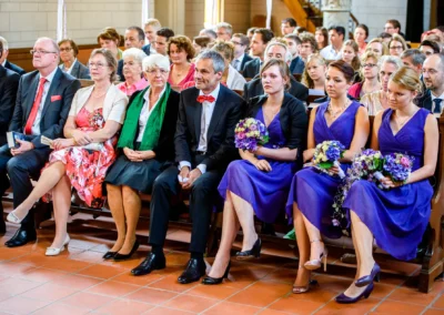 Gäste sitzen in einer Kirche, einige mit Blumensträußen, festlich gekleidet, violette Kleider heben sich hervor.