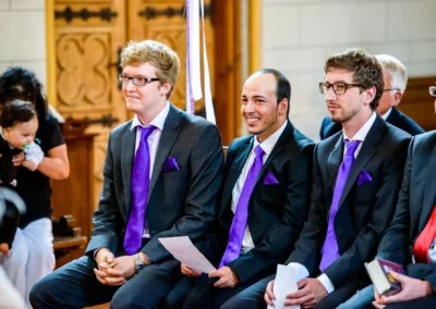 Mehrere Männer in Anzügen mit lila Krawatten sitzen auf einer Kirchenbank, einer lächelt, ein anderer hält Papier.