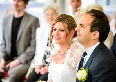 Braut und Bräutigam sitzen lächelnd nebeneinander, die Braut trägt ein weißes Kleid mit Spitzenjacke, Rose im Knopfloch des Bräutigams.