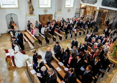 Brautpaar kniet in der Kirche, umgeben von stehenden Gästen in festlicher Kleidung, alle halten Kirchenhefte.