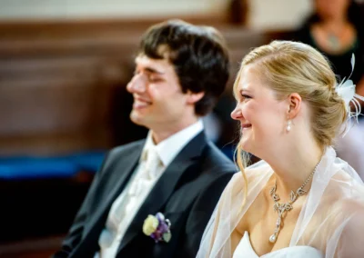 Ein Brautpaar sitzt lächelnd nebeneinander. Die Braut trägt Schmuck und ein weißes Kleid, der Bräutigam einen Anzug mit Blume.