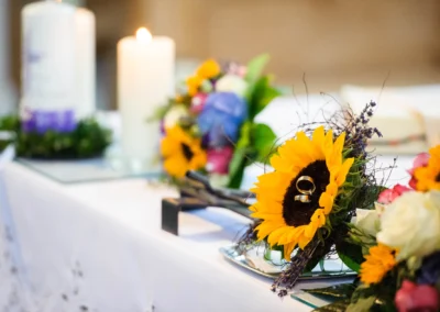 Ein Tisch mit Kerzen und einem Blumenarrangement aus Sonnenblumen, auf dem Eheringe liegen.