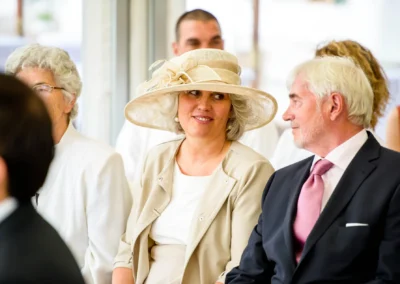 Ein elegantes Paar sitzt lächelnd in Hochzeitsgesellschaft, die Frau trägt einen großen Hut, beide sind in festlicher Kleidung.