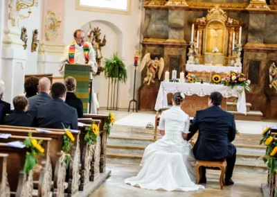 Ein Brautpaar kniet vor dem Altar in einer Kirche, während ein Geistlicher predigt. Bänke sind mit Sonnenblumen dekoriert.