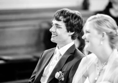 Ein Mann im Anzug und eine Frau lächelnd in einem weißen Kleid sitzen nebeneinander in einem Raum mit Holzbänken.