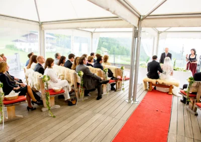 Hochzeitszeremonie in einem Zelt mit Bergpanorama, Gäste sitzen auf dekorierten Bänken, Brautpaar vor dem Trautisch.