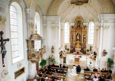 Helle Kirche mit verziertem Altar. Brautpaar sitzt vorne, Priester spricht. Gäste in Bänken, Barockelemente verzieren den Raum.