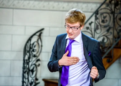 Ein Mann im Anzug mit lila Krawatte fasst sich an die Brust, im Hintergrund eine verzierte Treppe.