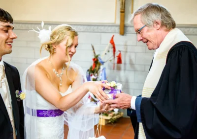 Braut und Bräutigam übergeben einem Mann in liturgischem Gewand einen kleinen, dekorierten Gegenstand in einer Kirche.