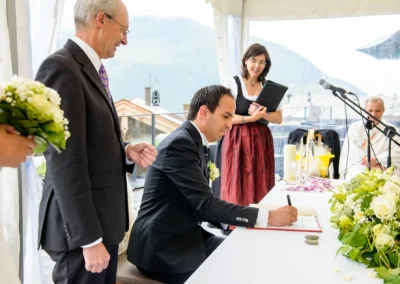 Ein Bräutigam unterschreibt das Traubuch, daneben ein Mann, eine Frau mit Mappe und ein Brautstrauß sichtbar.