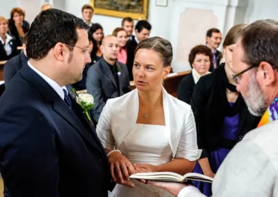 Ein Brautpaar tauscht Ringe in einer Kirche, umgeben von Gästen. Die Braut trägt ein weißes Kleid, der Bräutigam einen dunklen Anzug.