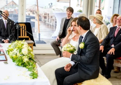 Brautpaar sitzt während der Trauung; Braut wischt Träne ab, hält Strauß, umgeben von Gästen. Elegantes, emotionsgeladenes Setting.