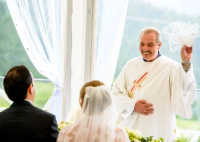 Ein lächelnder Priester in weißem Gewand führt eine Trauung durch; Braut und Bräutigam sitzen davor.
