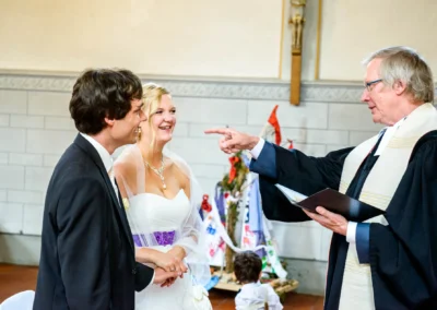 Ein Paar lacht, während ein Pastor im Talar mit Buch in der Hand etwas betont gestikuliert.