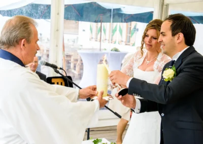 Brautpaar zündet Kerze mit Priester an; Braut im weißen Kleid, Bräutigam im dunklen Anzug, feiern Zeremonie in Zelthalle.