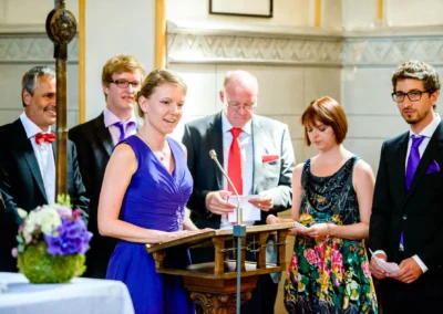 Eine Frau in lila Kleid spricht am Rednerpult, umgeben von elegant gekleideten Personen in einer Kirche.