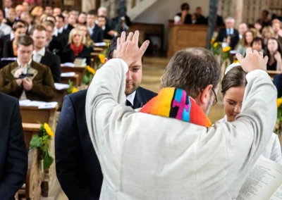 Ein Geistlicher segnet ein Brautpaar in einer Kirche, umgeben von Gästen in festlicher Kleidung.
