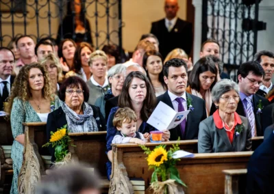 Gäste sitzen in Kirchenbänken mit Sonnenblumen, einige halten Programme. Eine Frau hält ein Kind auf dem Schoß.