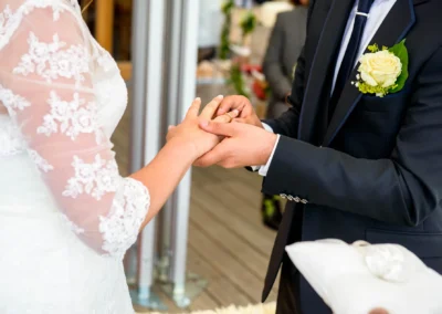 Ein Bräutigam steckt der Braut einen Ring an den Finger. Sie trägt ein weißes Kleid mit Spitzenärmeln.