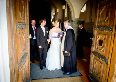 Das Brautpaar verlässt lachend die Kirche, begleitet von einem Geistlichen. Die Braut trägt ein weißes Kleid mit violettem Blumenstrauß.