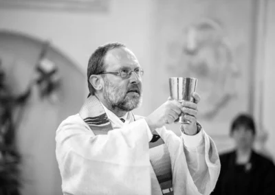 Ein Priester hält einen Kelch und trägt eine Stola. Hintergrund unscharf, leere Kirche sichtbar.