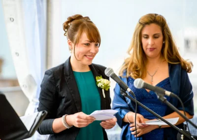 Zwei Frauen stehen lächelnd am Mikrofon, eine trägt einen Blazer mit Blumenschmuck, die andere ein blaues Kleid.