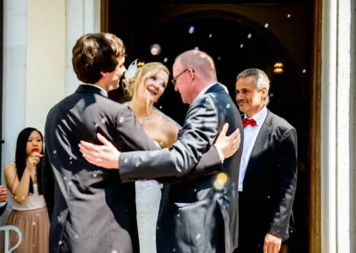 Das Brautpaar lacht glücklich in der Sonne, während Gäste draußen Seifenblasen pusten. Ein herzliches Umarmen.