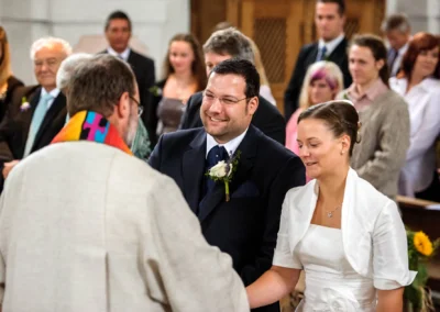 Ein Brautpaar steht fröhlich vor einem Geistlichen in der Kirche. Gäste im Hintergrund beobachten die Zeremonie.