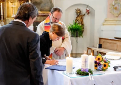 Eine Frau unterschreibt ein Dokument in der Kirche, ein Priester und ein Mann stehen daneben, Kerzen und Blumen zieren den Tisch.
