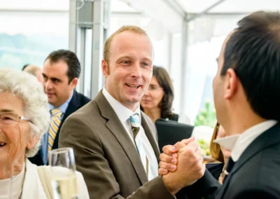 Mehrere Menschen in festlicher Kleidung, ein Mann lächelt und gibt Händedruck, eine ältere Frau lacht, Sektempfang.