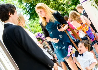 Ein Kind mit Seifenblasenpistole lacht, während Gäste in festlicher Kleidung plaudern und eine Braut einen Blumenstrauß hält.