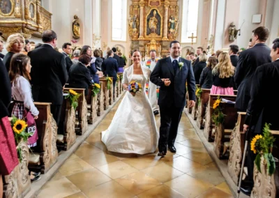 Brautpaar schreitet lächelnd durch Kirchenschiff, Braut trägt weißes Kleid, Bräutigam Anzug, fotografiert von einem Hochzeitsfotografen in Zorneding.