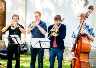 Eine vierköpfige Band mit Blechblasinstrumenten und Kontrabass spielt im Freien; alle tragen Hemden, zwei mit Notenständern.