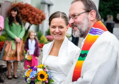 Die Braut in weißem Kleid mit buntem Blumenstrauß lächelt neben einem Mann mit buntem Schal. Im Hintergrund Gäste.