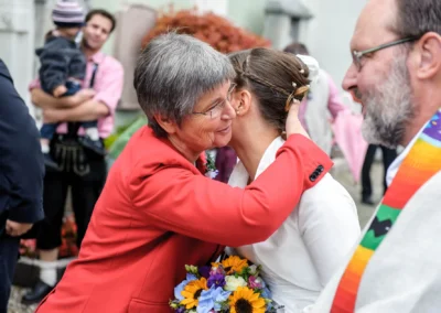 Zwei Personen umarmen sich herzlich; die Braut hält einen bunten Blumenstrauß. Im Hintergrund steht eine Person mit Kind.