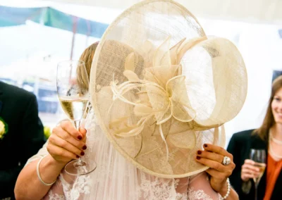 Eine Person in elegantem Kleid und großem Hut hält ein Sektglas, eine lächelnde Frau im Hintergrund.
