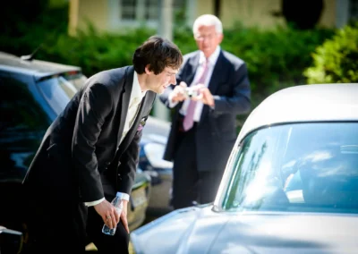Ein Mann im Anzug beugt sich lächelnd zu einem Auto; im Hintergrund fotografiert ein älterer Herr.