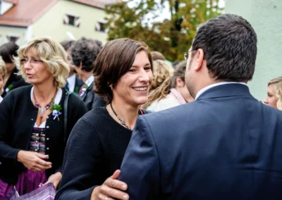 Zwei lächelnde Personen begrüßen sich bei einer Feier im Freien, umgeben von festlich gekleideten Gästen.