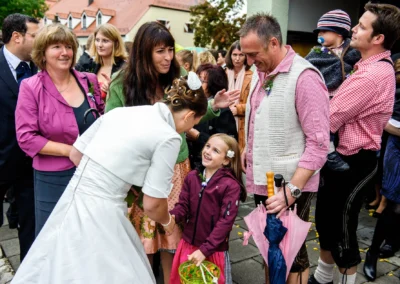 Eine Braut begrüßt ein lachendes Mädchen mit Korb, umgeben von festlich gekleideten Gästen.