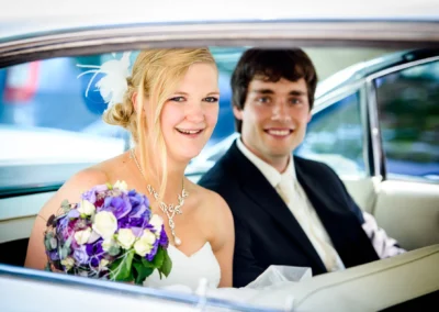 Ein lachendes Brautpaar sitzt im Auto, die Braut hält einen lila-weißen Blumenstrauß. Beide sind festlich gekleidet.