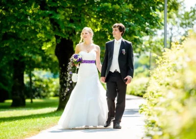 Ein Brautpaar spaziert auf einem sonnigen Weg, sie trägt ein weißes Kleid mit lila Blumenstrauß, er einen dunklen Anzug.