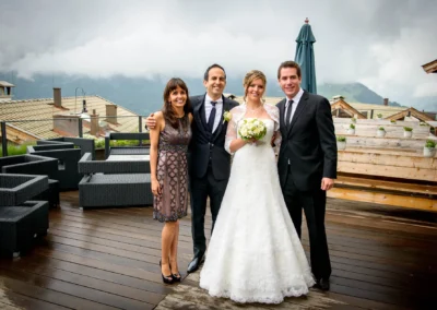 Das Brautpaar steht mit zwei Gästen auf einer Terrasse. Die Braut trägt ein weißes Kleid und hält einen Blumenstrauß.