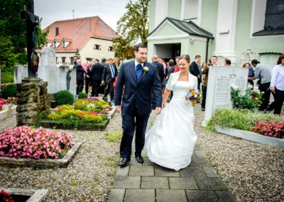 Das Brautpaar schreitet lächelnd Arm in Arm über einen Friedhof, umgeben von Gästen vor einer Kirche.