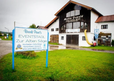 Ein altes Gebäude mit Holzschindeln, eventstadl Zur Alten Säge beschildert. Vorneweg ein grüner Rasen. Bewölkter Himmel.