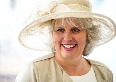 Eine lächelnde Frau in heller Kleidung und großem Hut, vor hellem Hintergrund.