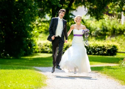 Ein Brautpaar läuft freudig einen Gartenweg entlang, die Braut trägt ein weißes Kleid mit lila Band, hält einen Blumenstrauß.